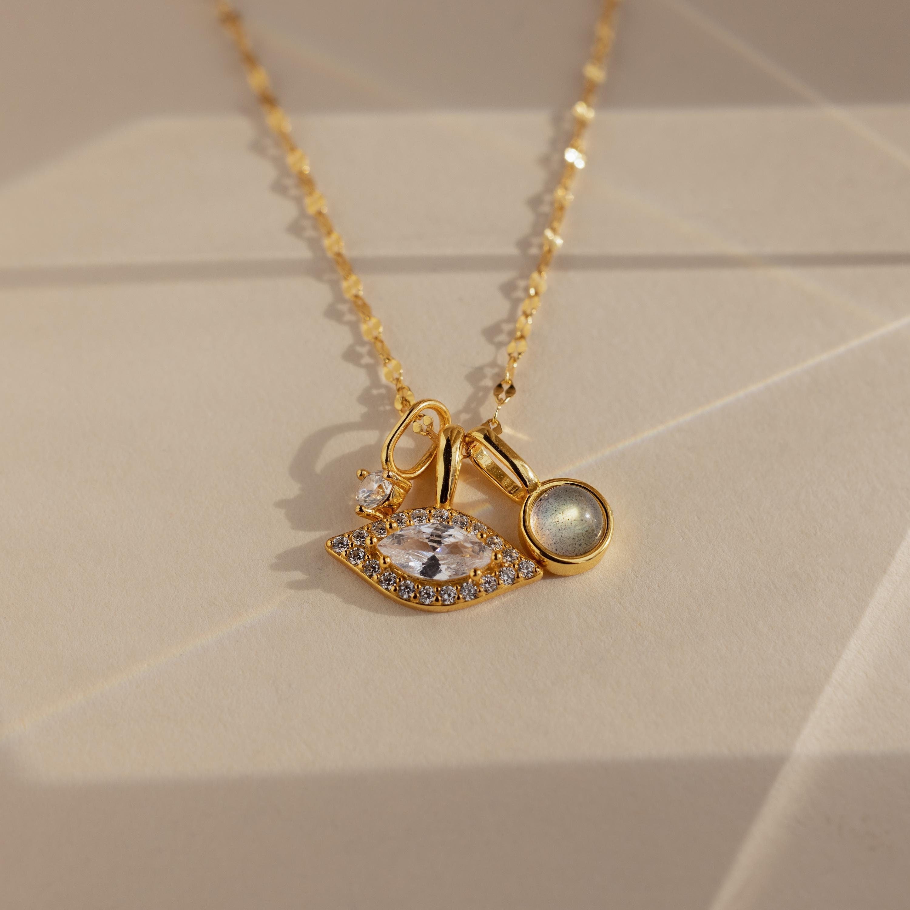 A gold chain necklace featuring a gold evil eye charm with a sparkling central cubic zirconia and encrusted smaller cubic zirconia stones, presented on a light background.
